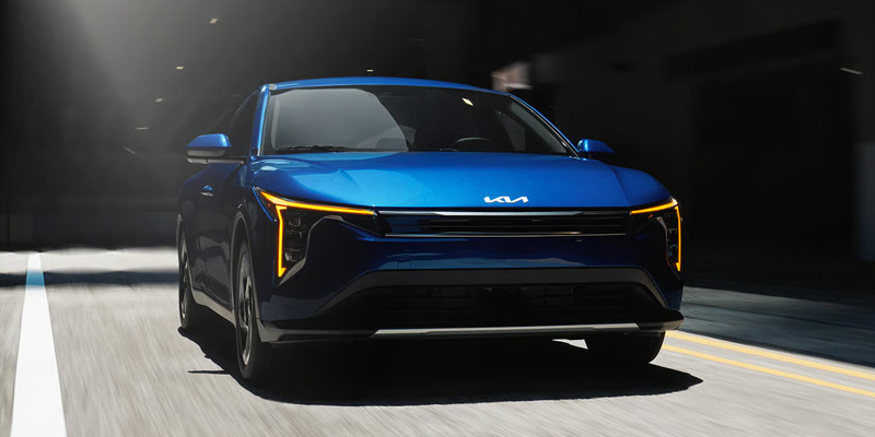 Blue Kia K4 sedan driving through a sunlit urban street, shown from the front.